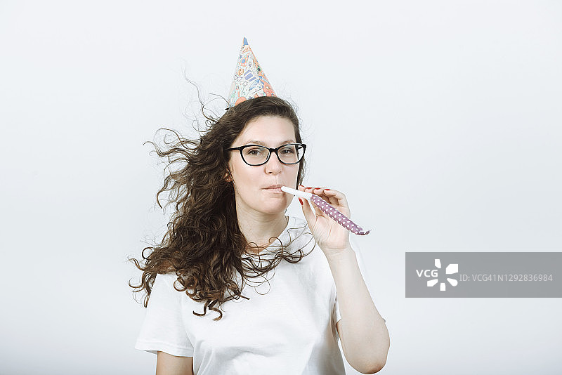 戴着生日帽吹口哨的快乐年轻女子，在灰色背景工作室里图片素材