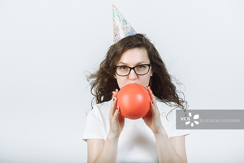 戴着眼镜和派对帽，在灰色背景工作室吹红色气球的女人图片素材