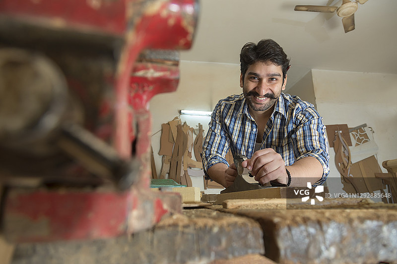 微笑的木匠用刮刀片平滑木头，前景有金属夹图片素材