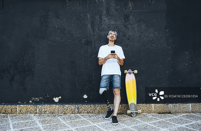 使用智能手机、带着假肢和滑板的笑容满面的年轻男子图片素材