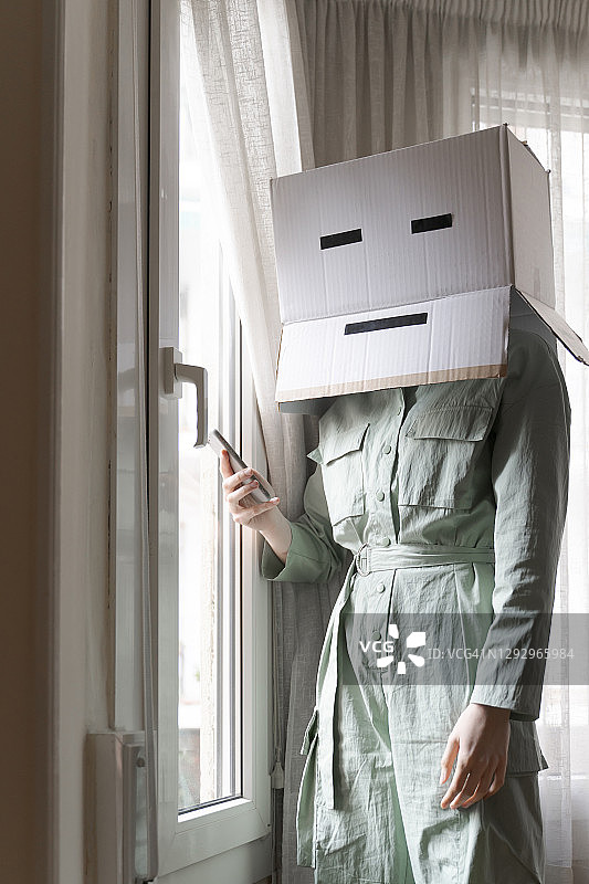 戴着纸板箱头，百无聊赖地从窗户往外看并使用智能手机的女人图片素材