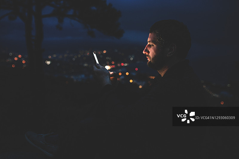 独自在夜间城市用手机：英俊的男人正在阅读消息或观看智能手机上的视频图片素材