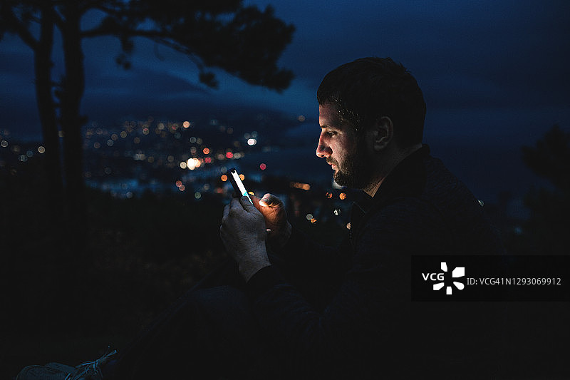 独自在夜间城市用手机：英俊的男人正在阅读消息或观看智能手机上的视频图片素材