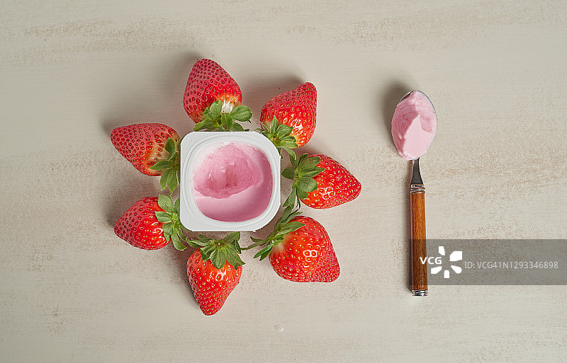 白色背景下，塑料酸奶杯里放着一勺红色酸奶，杯子周围有八颗带绿叶的草莓的俯视水平照片图片素材