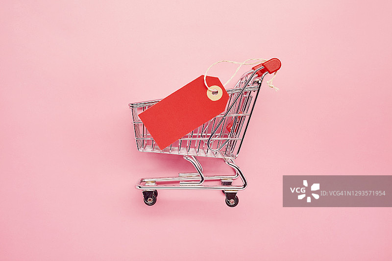 粉色背景上的带红色空白价格标签的小购物车静物，网上购物图片素材