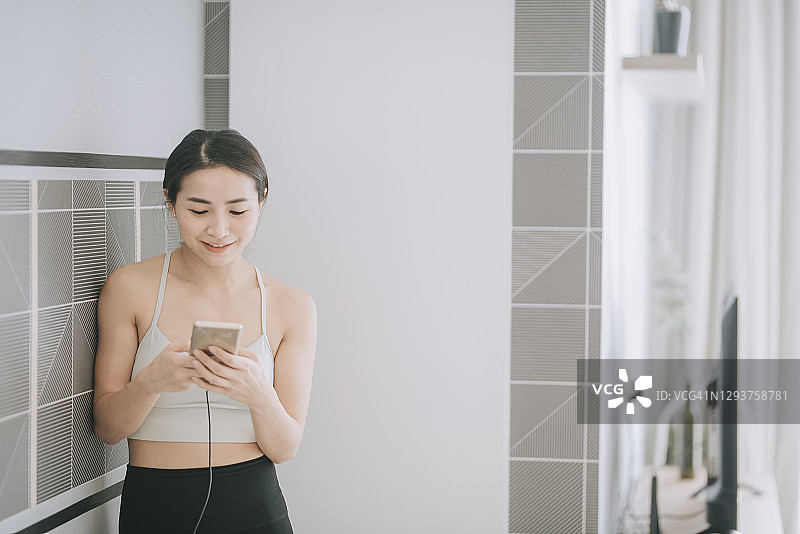 亚洲中国年轻女子靠墙休息，在客厅做瑜伽后查看智能手机图片素材