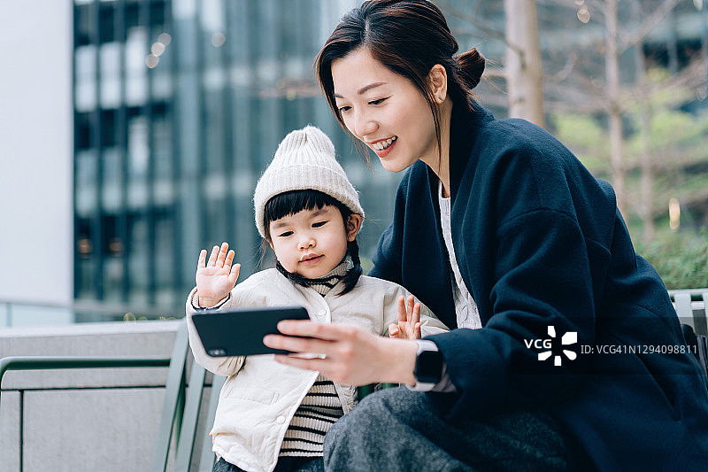 年轻的亚洲妈妈和可爱的女儿在户外用智能手机与家人朋友进行视频通话。她们坐在城市阳台的长椅上，背景是城市建筑。家庭和科技概念图片素材