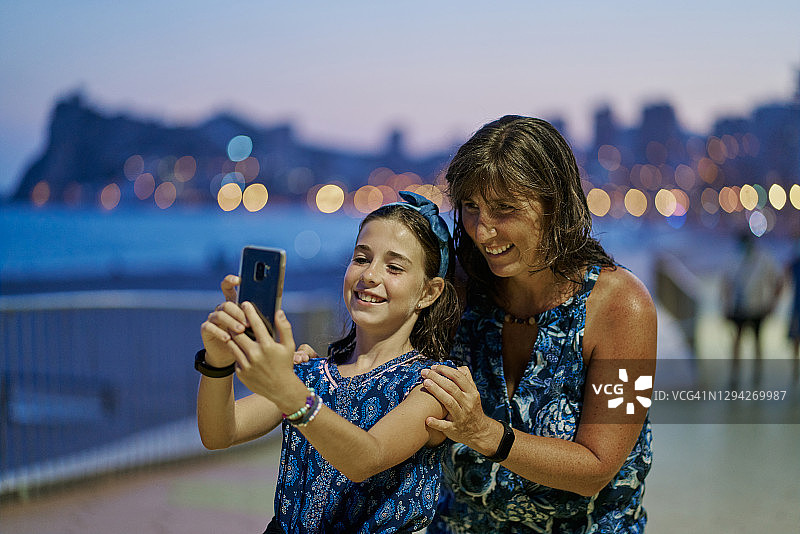 母亲和十几岁的女儿穿着蓝色连衣裙，晚上在海滩背景下用智能手机自拍。度假概念图片素材