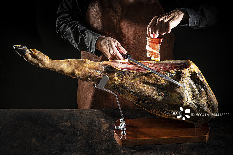 在阴暗背景下手持刀切割伊比利亚火腿的男人图片素材