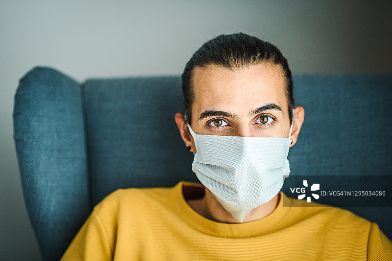 戴着医用防护口罩坐在椅子上看着镜头的年轻男人。室内照片。新冠病毒防护/隔离期间待在家中概念。图片素材