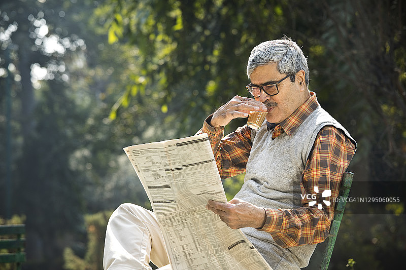 年长的男人在公园长椅上喝茶看报纸图片素材
