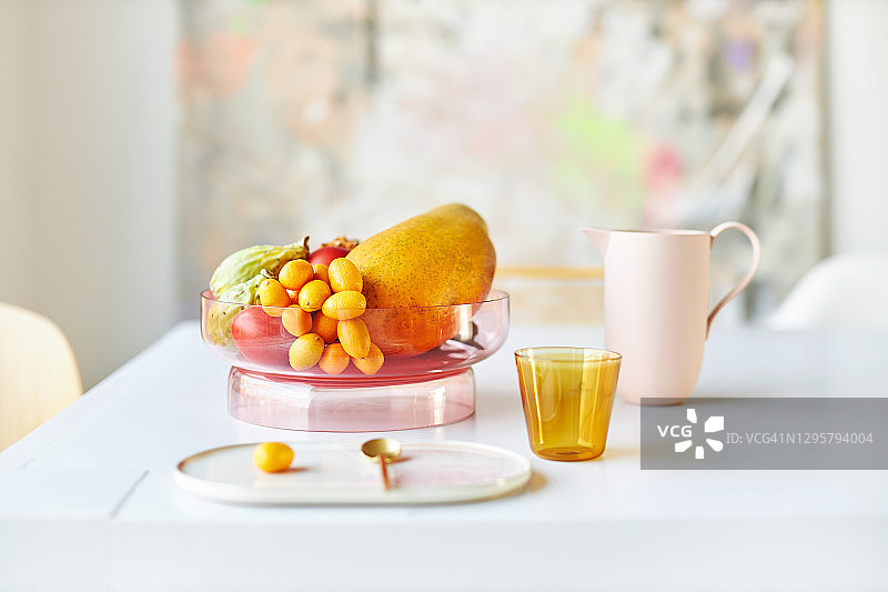 现代粉色玻璃果盘，盛满异国情调的水果，置于白色桌子上，旁边放着黄色水杯、设计盘和金色勺子，日光下拍摄图片素材