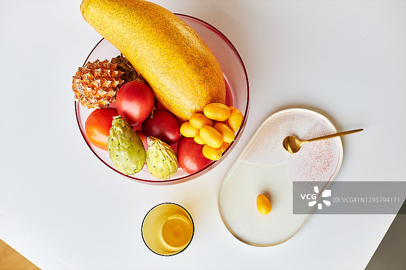 现代粉色玻璃果盘，盛满异国情调的水果，置于白色桌子上，旁边放着黄色水杯、设计盘和金色勺子，日光下拍摄图片素材
