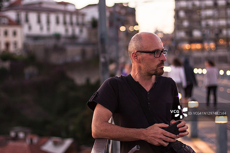 男人穿着黑色休闲装和眼镜，在日落时分站在波尔图路易一世大桥的栏杆上，手持智能手机图片素材