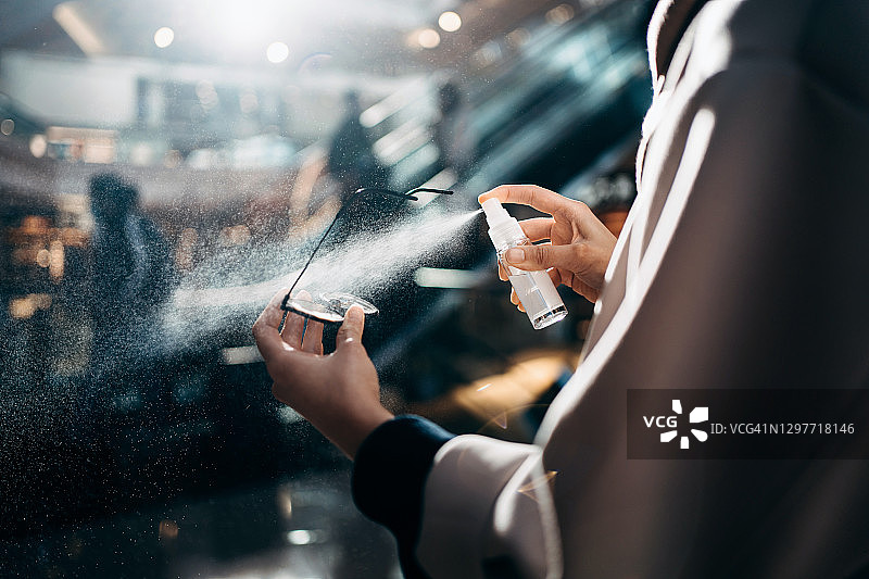 在购物中心购物时，年轻亚洲女性用消毒喷雾消毒眼镜，背景是阳光、自动扶梯和购物者图片素材