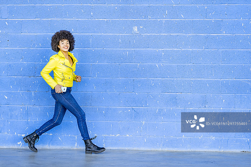 快乐的女人穿着黄色皮夹克靠在蓝色墙上图片素材