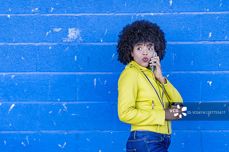 对着蓝墙，在电话中表现出惊讶反应的非洲裔年轻女子图片素材
