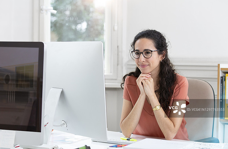 在办公室隔间里手托下巴工作的微笑女商人图片素材