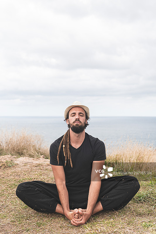 年轻男人在多云的天空下坐在田野上练习瑜伽，西班牙桑坦德图片素材