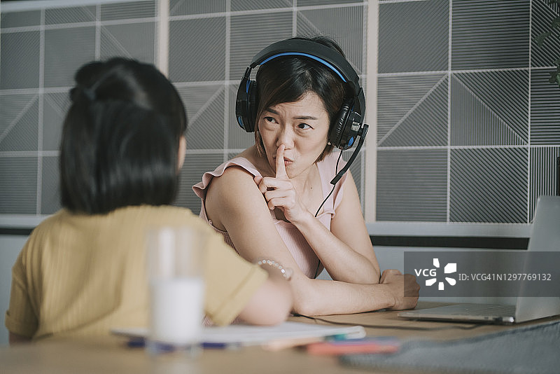 中国母亲在餐桌上用手指示意女儿保持安静，居家办公视频通话图片素材