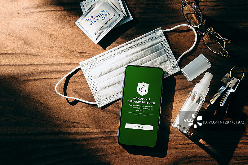疫情期间的日常必需品：外科口罩、消毒喷雾、酒精湿巾和智能手机。智能手机上的新冠病毒接触者追踪应用程序显示用户未感染。图片素材