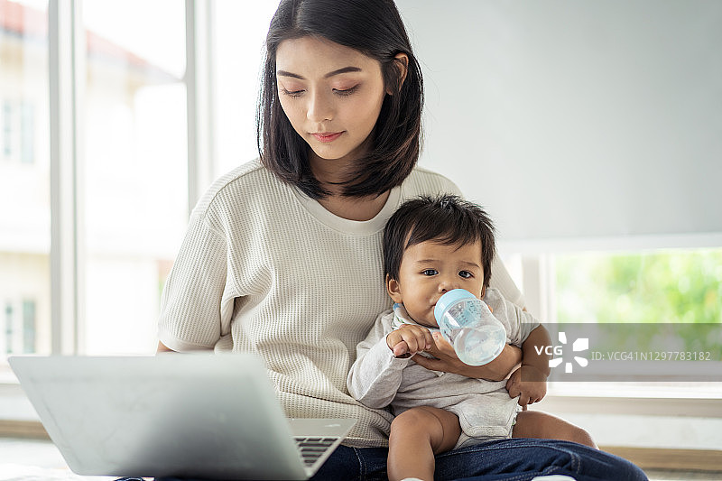 母亲在家用笔记本电脑工作时给宝宝喂奶瓶图片素材