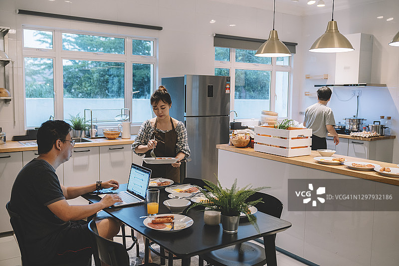 亚洲男人在家用笔记本工作，妻子在厨房准备早餐图片素材