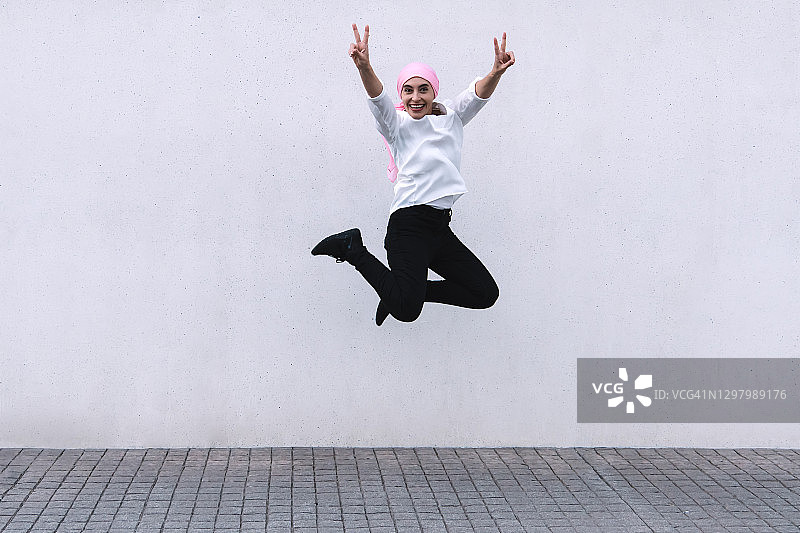 佩戴粉色围巾的女人，身穿黑色裤子和白色衬衫，双腿并拢跳跃，双手做出胜利手势图片素材