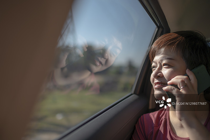坐在车内用手机通话的亚洲中年妇女，车窗上反映出电线和天空图片素材