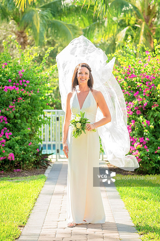 穿着婚纱站在花园里，全身，面纱在风中飘扬并拿着花束的新娘图片素材