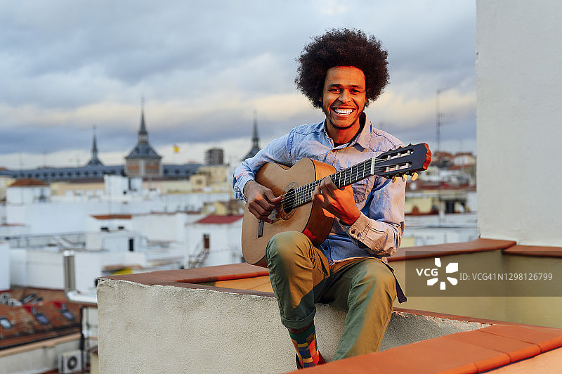 在露台上对着日落天空弹吉他的微笑青年图片素材