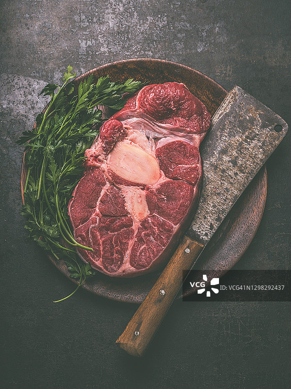 深色乡村背景木盘上配有新鲜烹饪香草和复古肉刀的生牛胫骨横切肉图片素材
