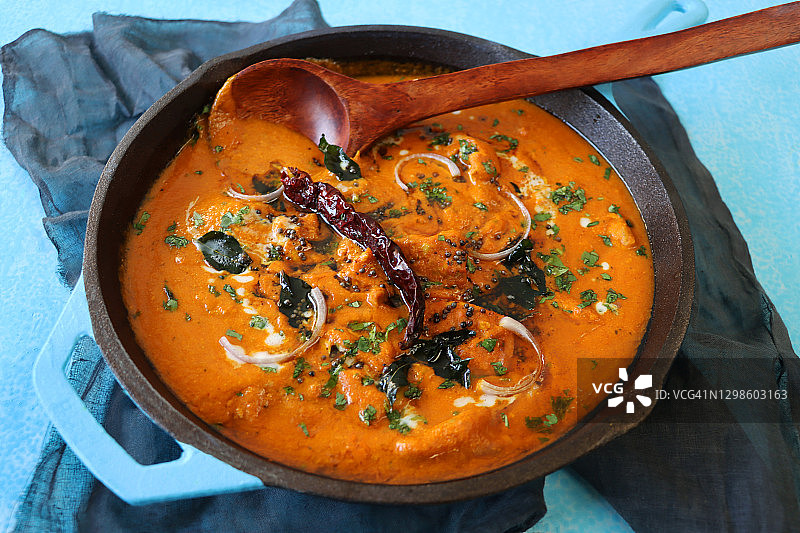 绿松石蓝色烹饪锅的特写图像，里面装满了印度黄油鸡肉/蒂卡咖喱，大块鸡胸肉在咖喱酱中，并配以红洋葱片和芥末籽装饰，高角度视图图片素材