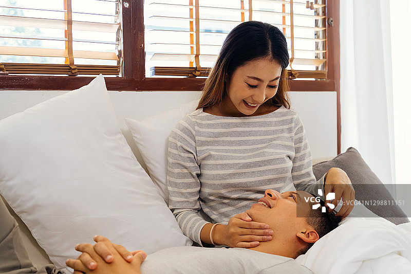卧室里幸福的亚洲年轻夫妇对视的场景，30多岁的成熟丈夫躺在妻子的腿上微笑，婚姻幸福生活图片素材