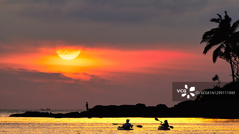 普吉岛岩石海滩日落时分，旅行者情侣划皮划艇，背景是椰子树和渔民图片素材