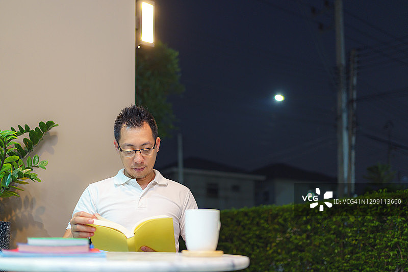 在泰国曼谷，一名亚洲男子在门廊下，开着灯，晚上看书，远处可见电塔图片素材