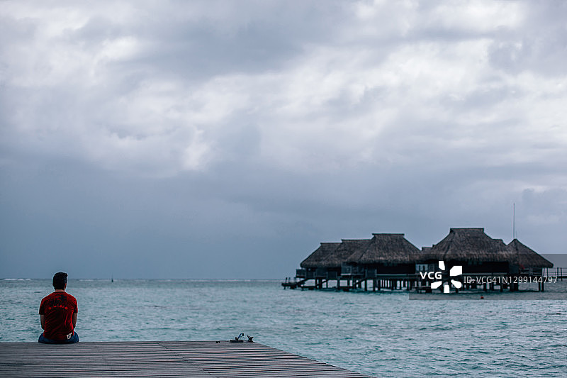 坐在码头上眺望太平洋的男人，背景是水上茅草屋顶的房屋，波利尼西亚莫雷亚岛图片素材