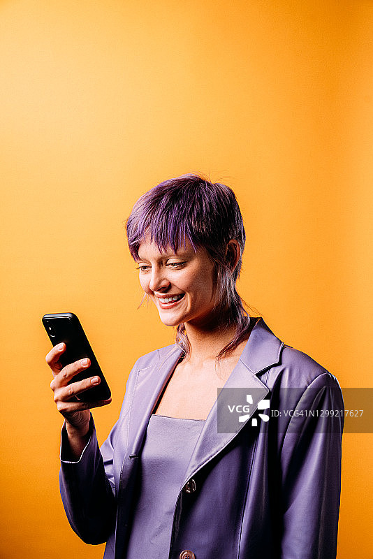 使用智能手机发短信、购物、手机银行或使用社交媒体的时尚自信美丽人士的影棚肖像，芥末黄色背景图片素材