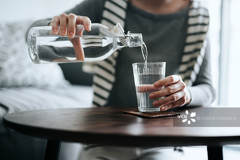 年轻亚洲女性在家中咖啡桌旁倒水特写：健康补水图片素材