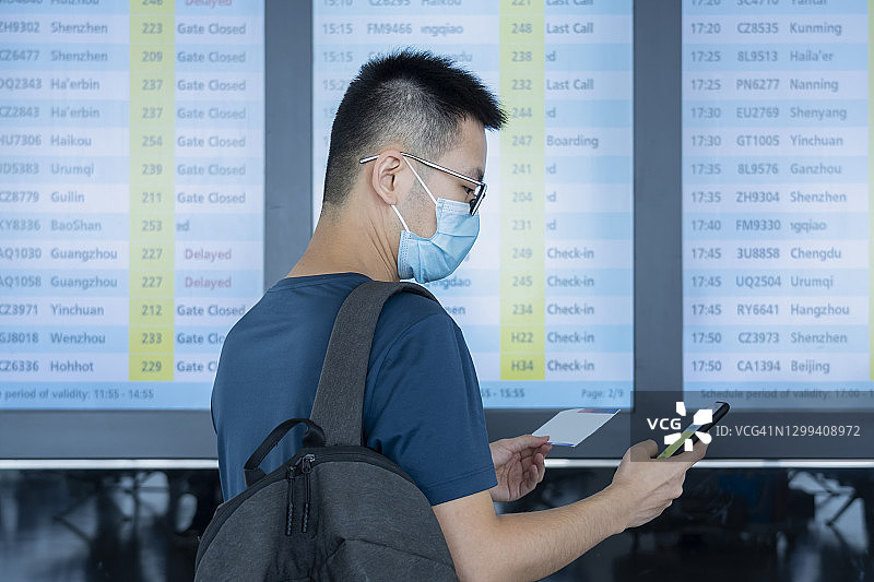 在机场航班信息屏幕前，一位戴着医用外科口罩、背着背包的亚洲男子正在用手机和机票查看登机信息图片素材