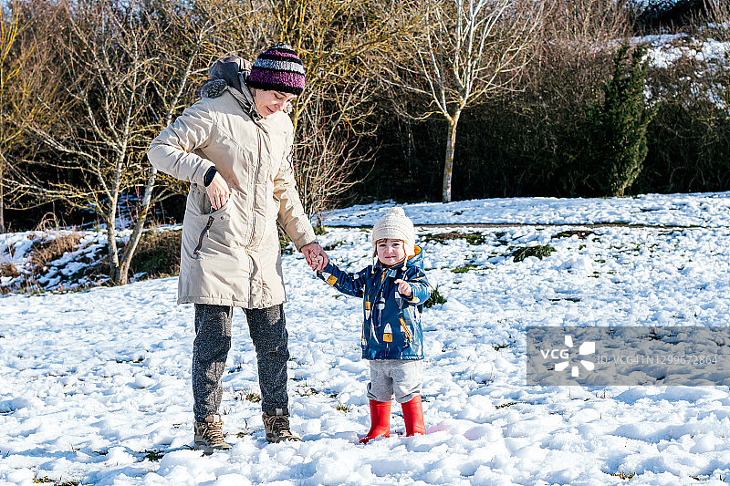小孩和母亲走在满是雪的田野上，小孩高兴地指着，穿着红色靴子和白色羊毛帽图片素材