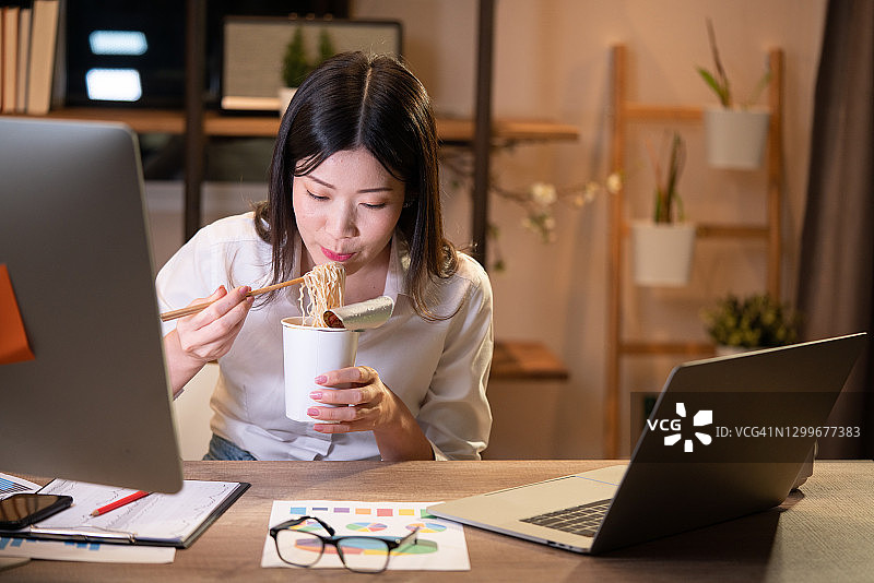 亚洲自由职业者：聪明商业女性在家中客厅晚上边吃方便面边用笔记本电脑工作图片素材
