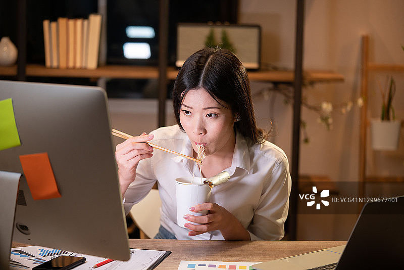 亚洲自由职业者：聪明商业女性在家中客厅晚上边吃方便面边用笔记本电脑工作图片素材