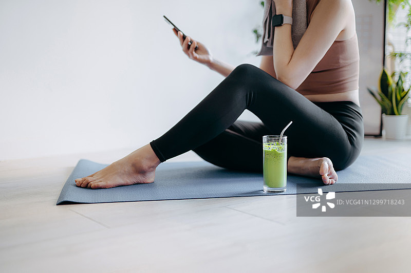 年轻亚洲运动女性在清晨在家锻炼后用健康绿色果汁补充水分并使用智能手机图片素材