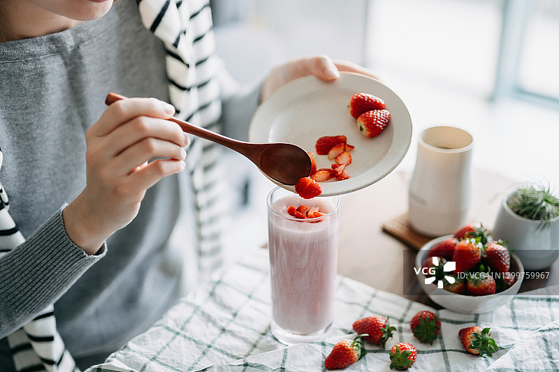 年轻的亚洲女性制作新鲜健康的草莓冰沙作为早餐，在顶部添加切碎的草莓作为装饰。健康饮食和健康的生活方式图片素材
