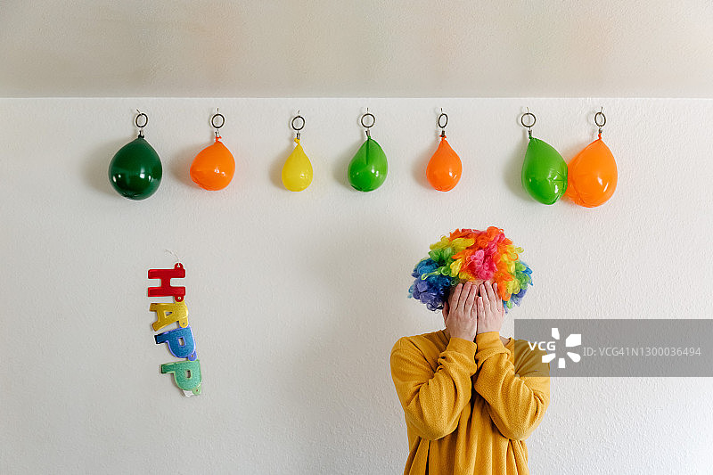 戴着小丑假发，站在瘪气球和破损的“生日快乐”标志旁的女人图片素材