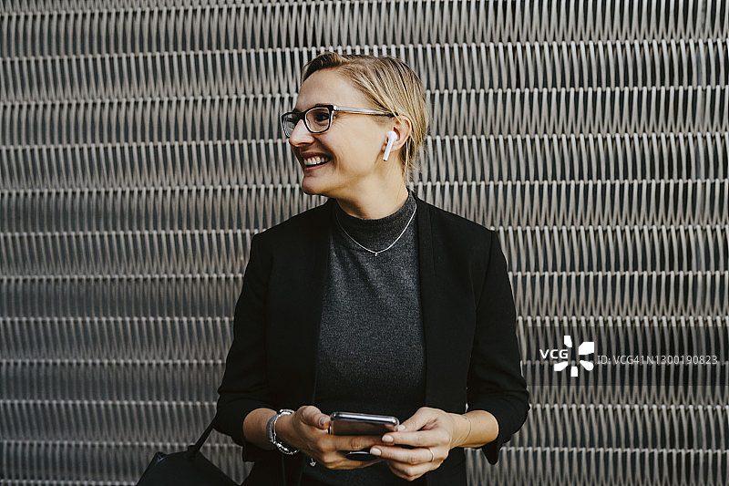 微笑的女商人戴着无线耳机听音乐，背景是金属格栅图片素材