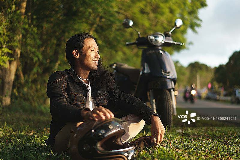 30多岁的东南亚男子坐在山路旁，绿色植物和经典摩托车相伴图片素材