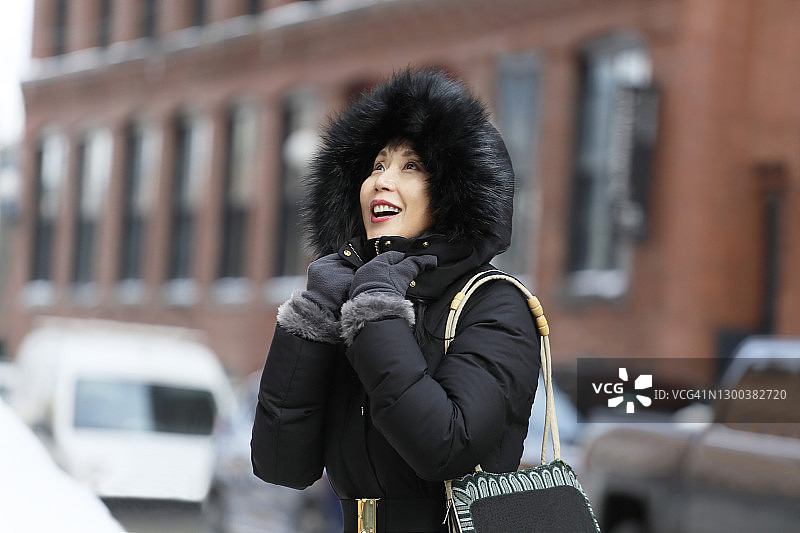一位身穿黑色毛皮连帽冬季外套的亚洲中年妇女站在城市工业砖砌建筑和汽车街道外，享受寒冷的天气。图片素材