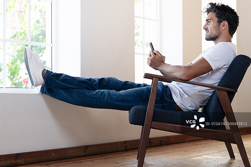 男人在家放松地坐在扶手椅上，用智能手机看着窗外图片素材